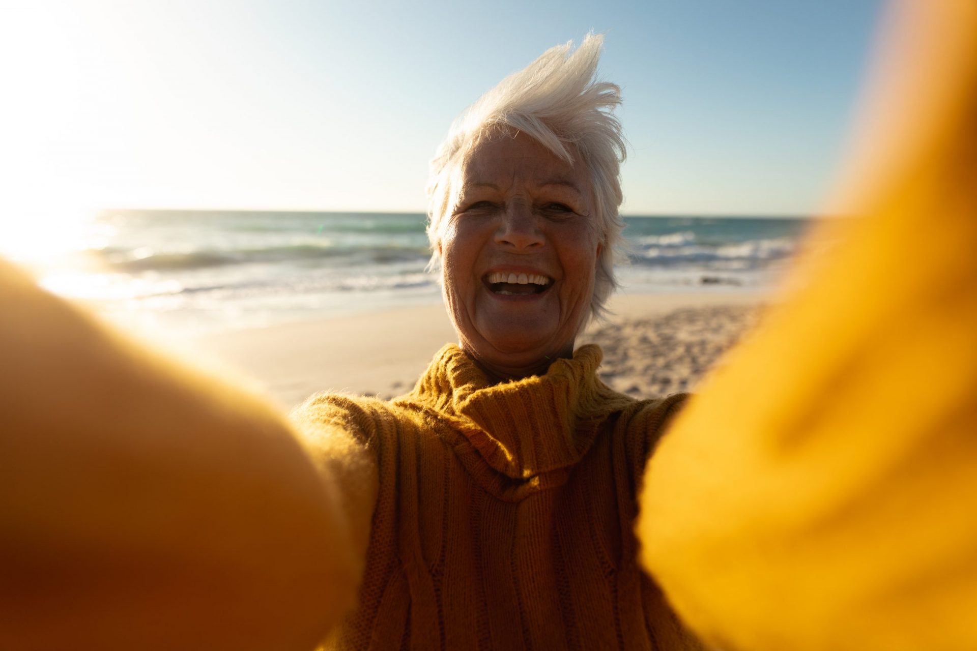 Front view of a senior Caucasian woman at the beach in the sun, smiling to camera and reaching out taking a selfie with a device out of shot
